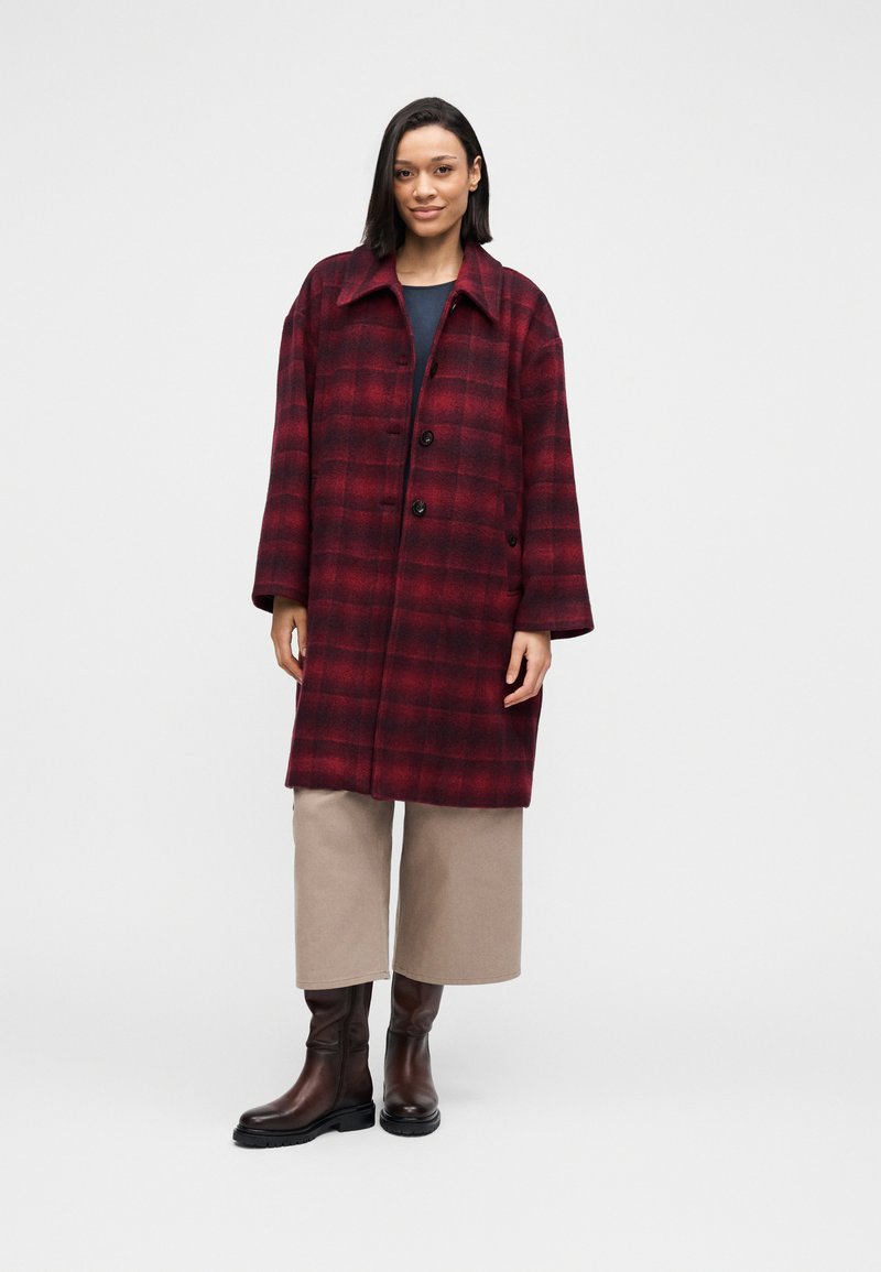 Red and black plaid oversized wool coat with two front buttons, broad collar, and side pockets, paired with beige cropped pants and brown boots.