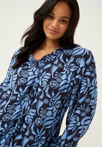 Bluse mit blauem Blumenmuster und marineblauem Hintergrund, mit rüschigem Ausschnitt und strukturierter Oberfläche. Lange Ärmel und lässiger Schnitt.
