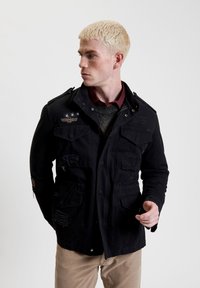 Zwarte katoenen militaire stijljack met meerdere zakken, versleten details en een drukknoopsluiting. Beschikt over een embleem en decoratieve stiksels.