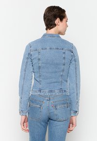 Ljusblå denimjacka med en figurnära design, horisontella och vertikala sömmar, samt en klassisk krage, sedd från baksidan.