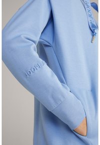 Sudadera azul claro con tejido texturizado, que incluye una capucha, un logo en relieve en la manga y bolsillos laterales. Diseño sencillo y casual.