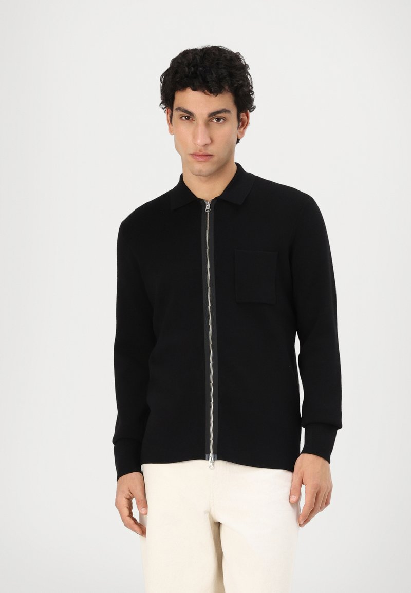 Maglione nero con zip, colletto, maniche lunghe e taschino sul petto, realizzato in un tessuto liscio e texturizzato.