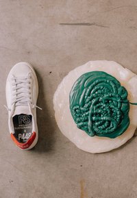 Baskets blanches avec un détail orange au talon à côté d'une assiette ronde tenant de la peinture verte épaisse sur un sol en béton gris.