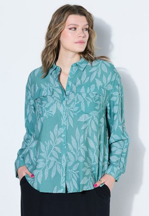 Mujer con camisa de botones color verde azulado con patrones de hojas blancas, manos en los bolsillos, mirando hacia un lado sobre un fondo claro y liso.