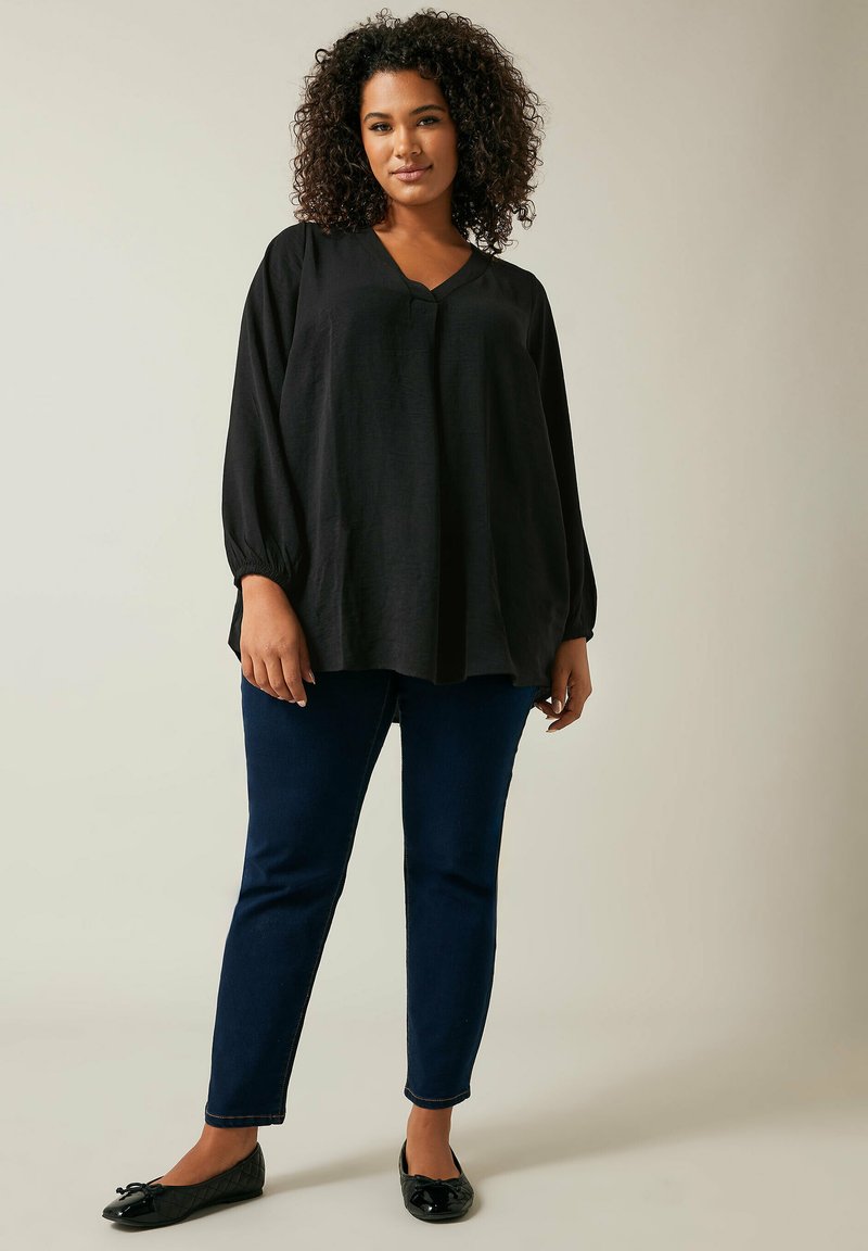 Schwarze Bluse mit langen Ärmeln und V-Ausschnitt sowie gerafften Manschetten, kombiniert mit dunkelblauen Slim-Fit-Jeans und schwarzen Ballettschuhen. Glatte Textur.