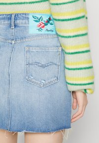 Džínová sukně světle modré barvy s třepeným lemem, obsahující zadní kapsy a vyšívanou nášivku s růžovým designem na modrém podkladu.