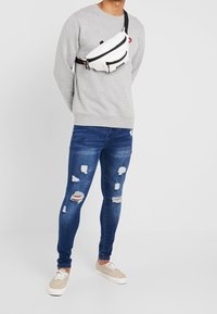 Sudadera gris, jeans azules desgastados con rasgaduras, zapatillas beige y una riñonera blanca con detalles en negro en el cierre y correa ajustable.