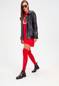 Chaqueta de motociclista de cuero negro sobre un vestido rojo con un logo blanco, combinada con medias altas rojas y botas negras hasta el tobillo.