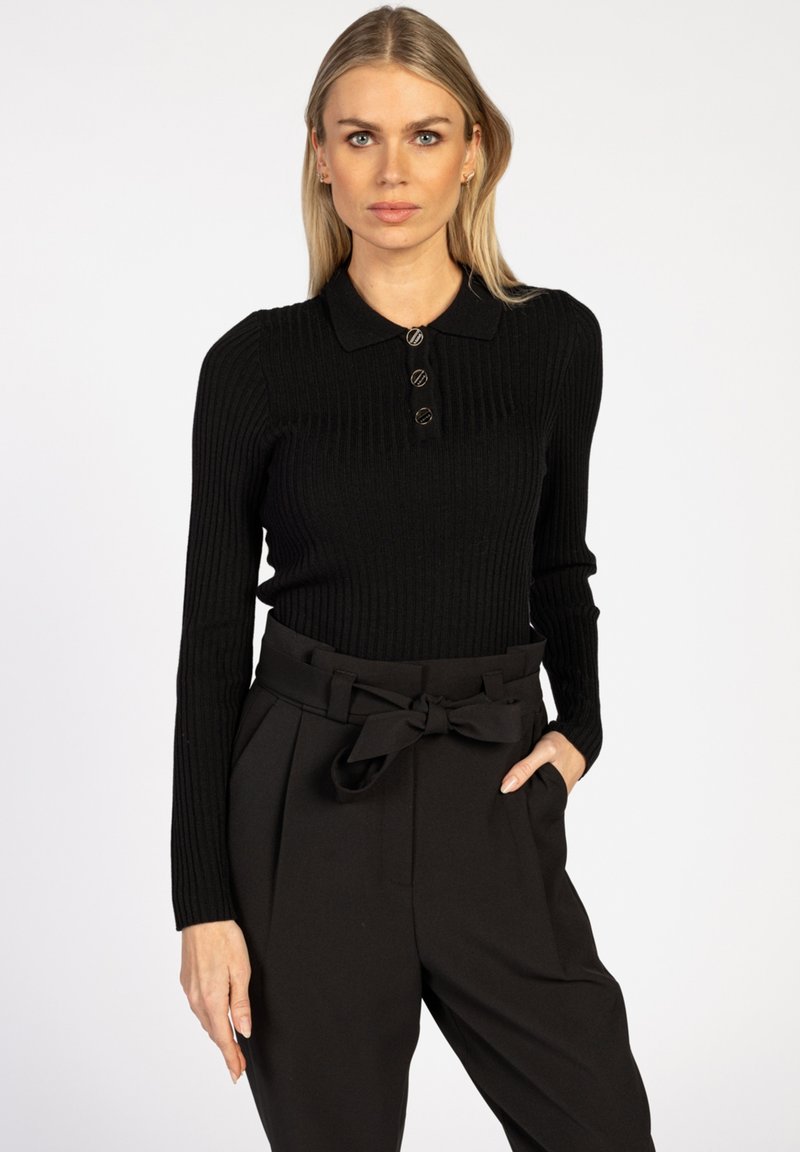 Blonde vrouw in een zwarte geribbelde lange mouwen polo trui en hoge taille zwarte broek met een gestrikte riem, staande met één hand in de zak.