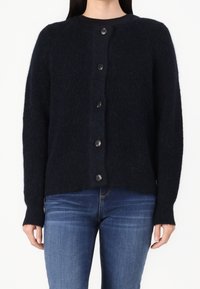 Kvinna med en svart fluffig knappad cardigan över blå jeans, stående mot en enkel vit bakgrund.