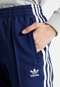 Marineblauwe sportbroek van een gestructureerde stof, met witte zijstrepen en een geborduurd Adidas-logo. Elastische tailleband met zijzakken.