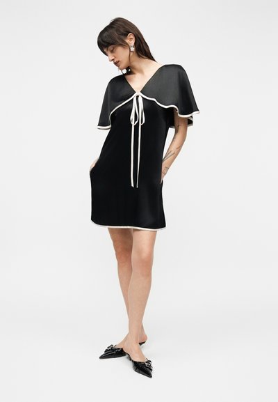 Γυναίκα που φοράει μαύρο σατινέ μίνι φόρεμα με λευκό τελείωμα και λεπτομέρεια μανδύα, συνδυασμένο με μαύρα μυτερά παπούτσια, στέκεται μπροστά σε λευκό φόντο.