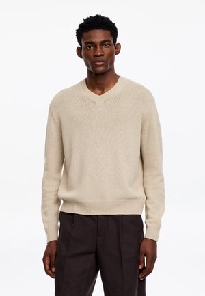 Homme portant un pull beige à col en V et un pantalon marron foncé, debout devant un fond blanc uni.