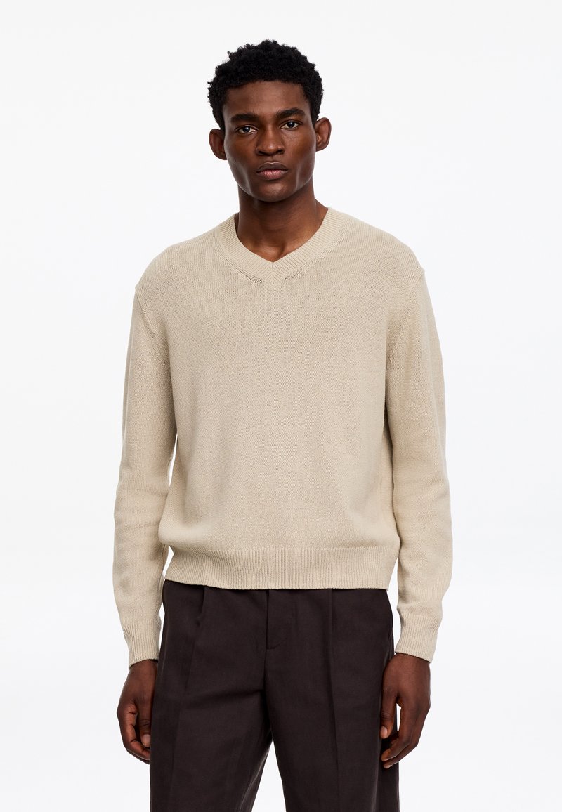 Homme portant un pull beige à col en V et un pantalon marron foncé, debout devant un fond blanc uni.