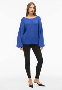Suéter azul texturizado con un escote amplio y mangas oversized, combinado con jeans pitillo negros y tacones de punta negros.