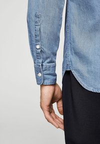 Ärmel eines Jeanshemds in Hellblau, mit geknöpften Manschetten und abgerundetem Saum. Glatte Textur mit sichtbaren Nahtdetails.