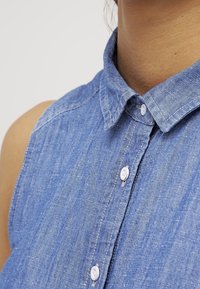 Ljusblå denimärmlös skjorta med en vikbar krage och knäppning. Har synlig sömnad och en texturerad tyg.