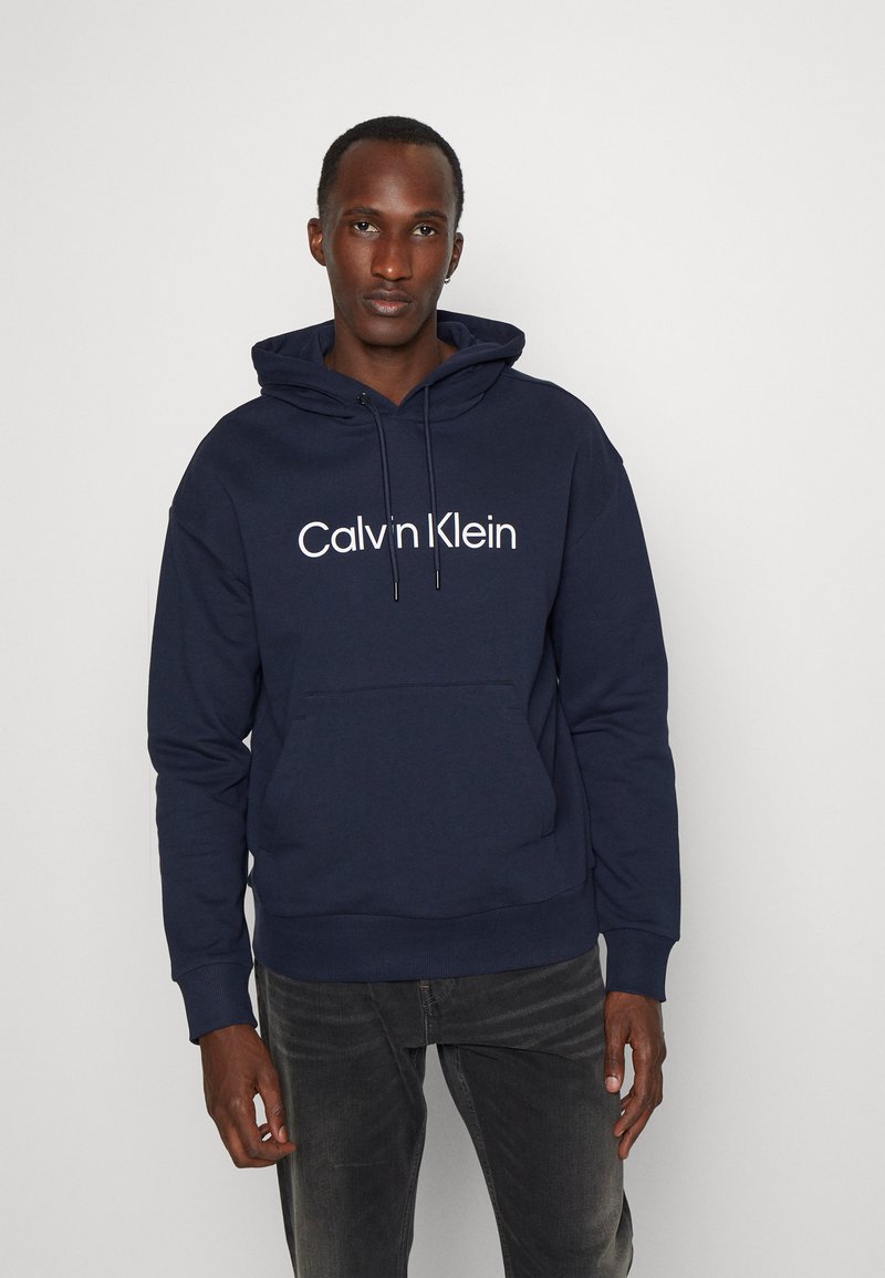 Navy blue hoodie with a front pocket, drawstrings, and a white "Calvin Klein" logo. Made from soft fabric with ribbed cuffs.