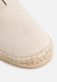 Zapatilla de lona beige con puntera redondeada, que cuenta con una suela de cuerda de yute y un intrincado bordado a lo largo del borde.