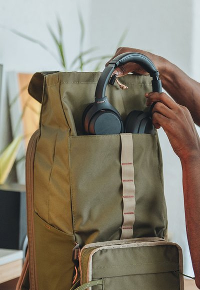 Händer som lägger svarta Sony-hörlurar i framfickan på en olivgrön ryggsäck inomhus, med suddiga växter i bakgrunden.