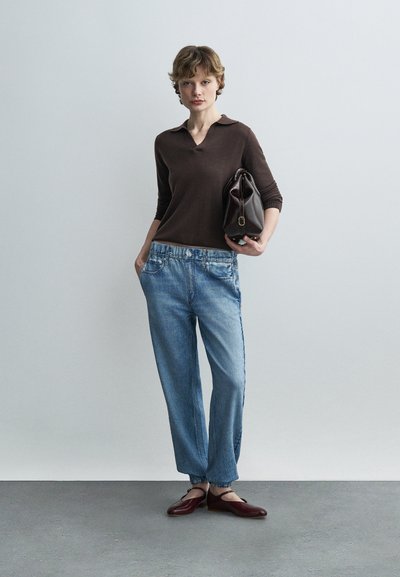 Νεαρό άτομο στέκεται μπροστά σε απλό φόντο φορώντας καφέ μπλούζα, φαρδιά μπλε τζιν, μπορντό φλατ παπούτσια και κρατώντας μια σκούρα καφέ τσάντα κάτω από το χέρι του.