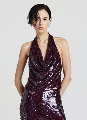 Frau mit nassem, kurzem, dunklem Haar, die ein ärmelloses, tiefpurpurnes, mit Pailletten besetztes Top und passende Hose vor einem schlichten, hellen Hintergrund trägt.