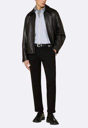 Giacca nera in pelle con cerniera sopra una camicia a righe azzurre chiusa con bottoni, abbinata a pantaloni neri e scarpe eleganti nere. Dettaglio fibbia della cintura in argento.