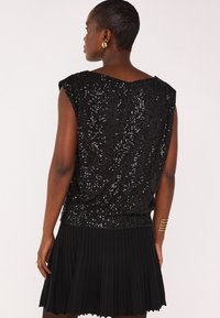 Top nero senza maniche realizzato in tessuto di paillettes, con una vestibilità ampia e orlo a coste. Abbinato a una gonna nera plissettata per creare contrasto.