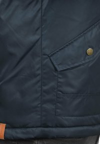 Giacca blu scuro con tasca a pressione e etichetta in pelle marrone vicino all'orlo, indossata su pantaloni scuri.