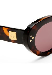 Κοντινό πλάνο γυαλιών ηλίου οβάλ σχήματος με μοτίβο χελώνας, μοβ φακούς και χρυσά διακοσμητικά καρφιά, καθώς και μικρό τετράγωνο χρυσό έμβλημα στο μπράτσο.