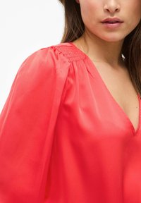 Blusa de seda coral con escote en V profundo, mangas abullonadas, detalles fruncidos en los hombros y textura suave. Enfoque en la parte superior del cuerpo.