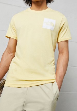 Person trägt ein blassgelbes kurzärmeliges T-Shirt mit The North Face Logo auf der Brust und hellfarbige Hosen, die Hände in den Taschen.