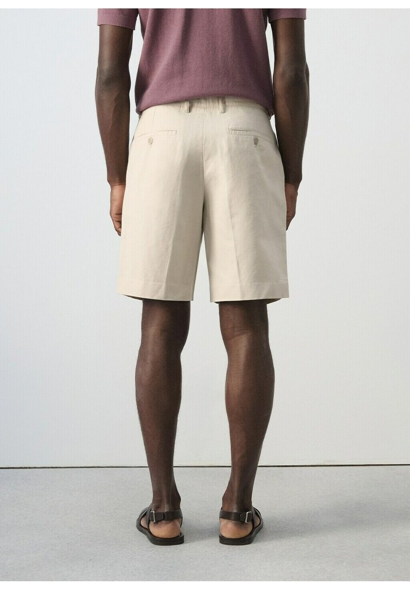 Homme debout de dos, portant un short beige mi-genou, une chemise mauve à manches courtes et des sandales noires avec des brides autour de la cheville.