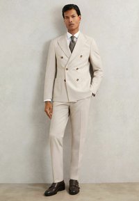 Bézs kétsoros zakó arany gombokkal, testhezálló szabással, bevágott hajtókkal, és egyenes szabású nadrággal, barna bőrcipővel és nyakkendővel párosítva.