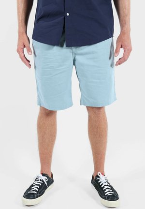 Homme portant un short bleu clair, des baskets noires avec des lacets blancs, et une chemise bleu marine à boutons, debout sur un fond blanc.