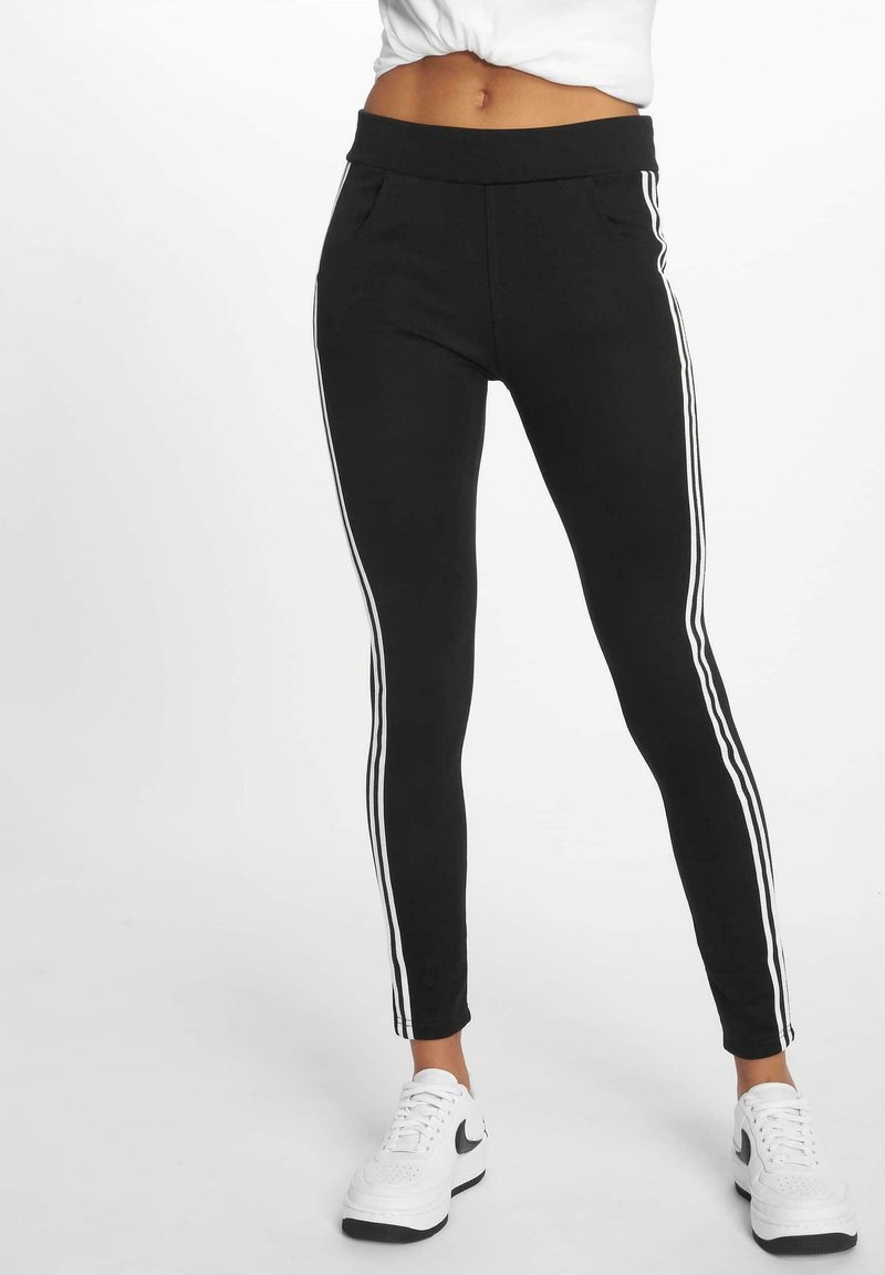 Leggings noirs avec une taille haute, comportant trois rayures blanches verticales le long des côtés extérieurs et deux poches à l'avant. Texture lisse.