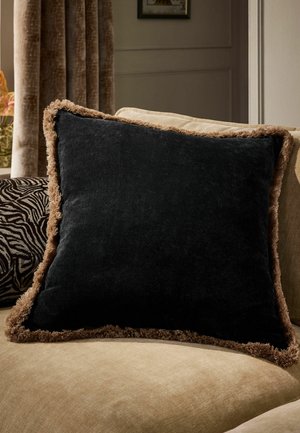 Quadratisches schwarzes Samtkissen mit braunem Fransenbesatz auf einem beigen Sofa in einem sanft beleuchteten Wohnzimmer.