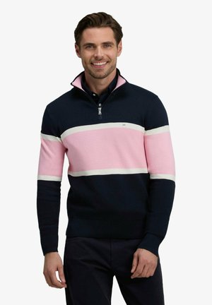 Homme souriant portant un pull à fermeture éclair quarter-zip rayé bleu marine et rose et un pantalon foncé, debout devant un fond uni.