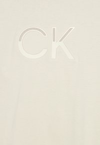 Lyse beige bomulds t-shirt med hævet hvid CK-tryk. Glat tekstur med en klassisk rund hals og korte ærmer.