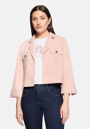 Kobieta w różowej krótkiej kurtce z kieszeniami z klapkami, nałożona na białą koszulkę z grafiką kółka i krzyżyka oraz ciemnoniebieskie, wysokie spodnie jeansowe.