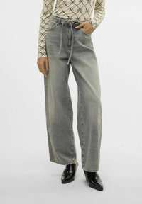 Ženske hlače iz sivih širokih jeans s pasom na vezanje, s pottonjem teksturo in dvema sprednjima žepoma, v kombinaciji s črnimi čevlji s peto.