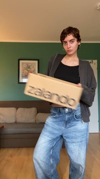 Kartono dėžė su juodu tekstu "zalando", laikoma žmogaus, apsirengusio tamsia sportine striuke ir šviesiais laisvais džinsais, gyvenamojoje patalpoje.