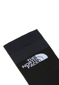 Calcetines negros con un puño gris oscuro. Presentan un logo blanco con el texto "THE NORTH FACE" y un diseño de montaña curvado. Textura acanalada en la parte superior.