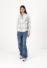 Camisa xadrez em branco, cinza e azul com dois bolsos no peito e fecho por botões; combinada com jeans flare azul claro e ténis cor de rosa.