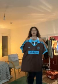 Καφέ κοντομάνικη φανέλα με ανοιχτό μπλε λεπτομέρειες και γιακά, με τρεις λωρίδες στα μανίκια και το λογότυπο "adidas" στο μπροστινό μέρος.