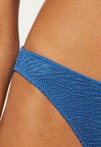 Niebieskie majtki bikini z teksturowanym, falistym wzorem dzianiny. Materiał jest elastyczny, z wąskim bocznym pasem odsłaniającym gładką skórę.