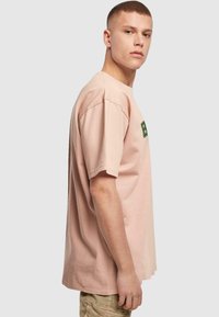 Lichtroze katoenen t-shirt met korte mouwen, een ontspannen pasvorm en een groen grafisch logo op de borst. Gladde textuur met minimale details.