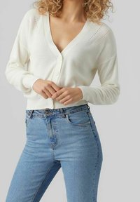 Witte gebreide cardigan met een V-hals, knoopsluiting aan de voorkant, een gestructureerd breipatroon en lange mouwen, gecombineerd met lichtblauwe, high-waisted jeans.