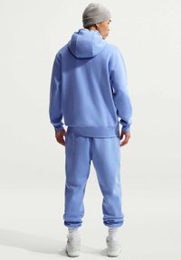 Hoodie bleu clair et joggers assortis en matière douce, avec une coupe décontractée et des poignets côtelés, portés avec un bonnet gris et des baskets blanches.