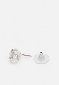 Cercei stud în formă de nod din argint, cu un finisaj texturat. Include un suport transparent pentru o purtare sigură. Design simplu, minimalism.